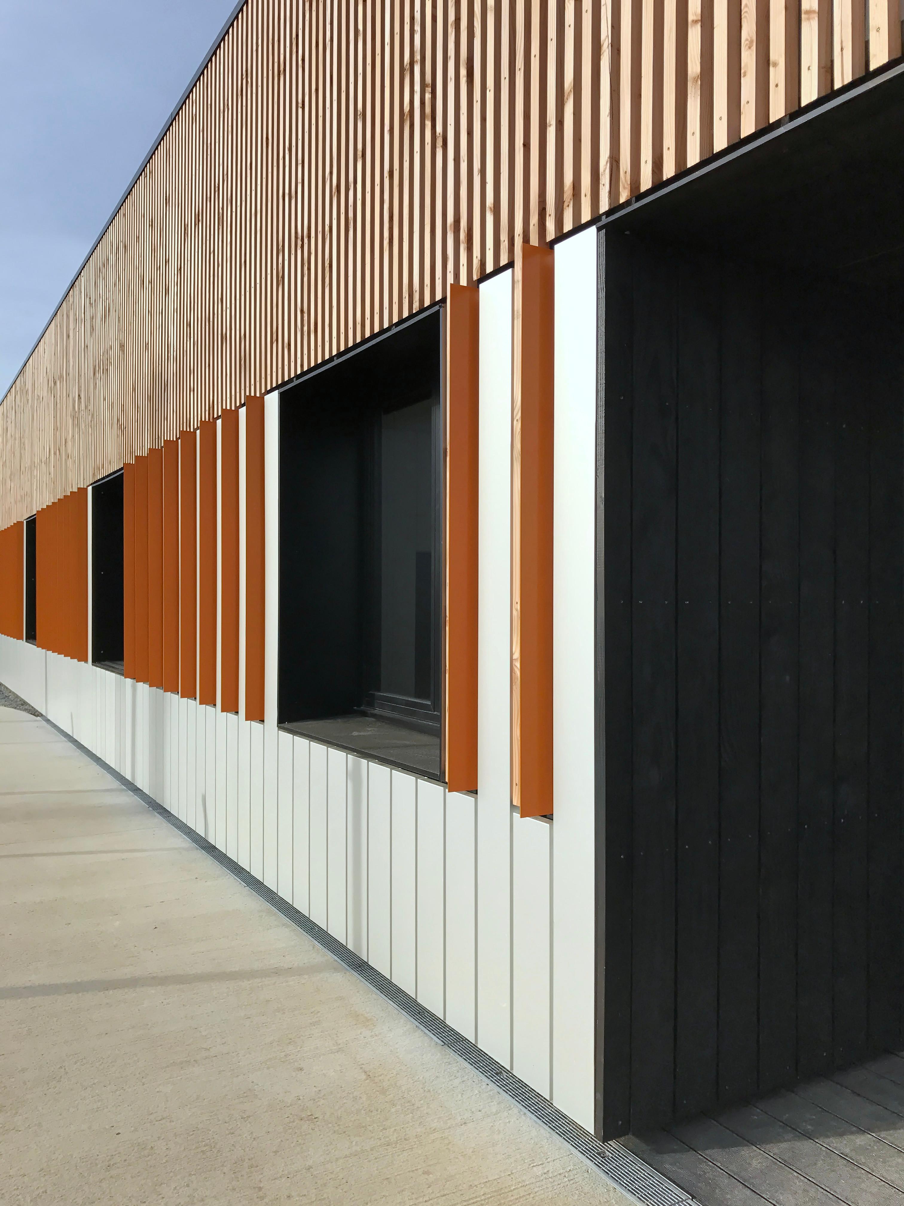 Photographie de la façade Sud. Cassettes aluminium brut, bardage bois & équerres en aluminium laquées orange.